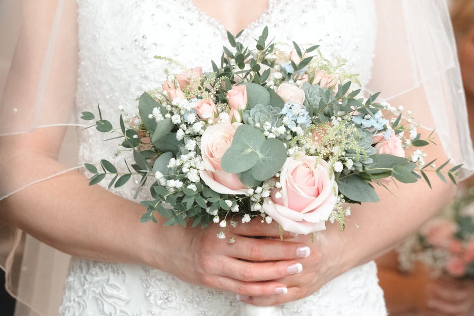 A bride in a white dress holding a floral bouquet arrangement featuring pale pink roses, eucalyptus leaves, and small white filler flowers, with the flowers arranged in a rounded cluster. The bouquet 