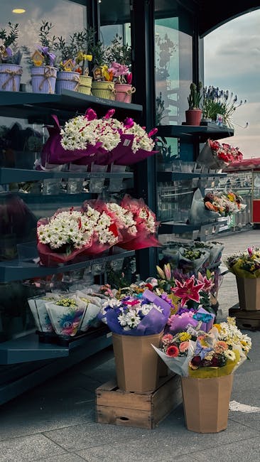 A display of fresh floral arrangements on dark grey shelves inside a flower shop, featuring bouquets wrapped in pink, purple, and brown paper, with a variety of flowers including white daisies, pink l
