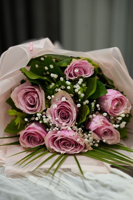 A bouquet of nine pink roses with delicate, tightly rolled petals, arranged with lush green leaves and small white baby's breath flowers. The flowers are wrapped in elegant white translucent paper wit